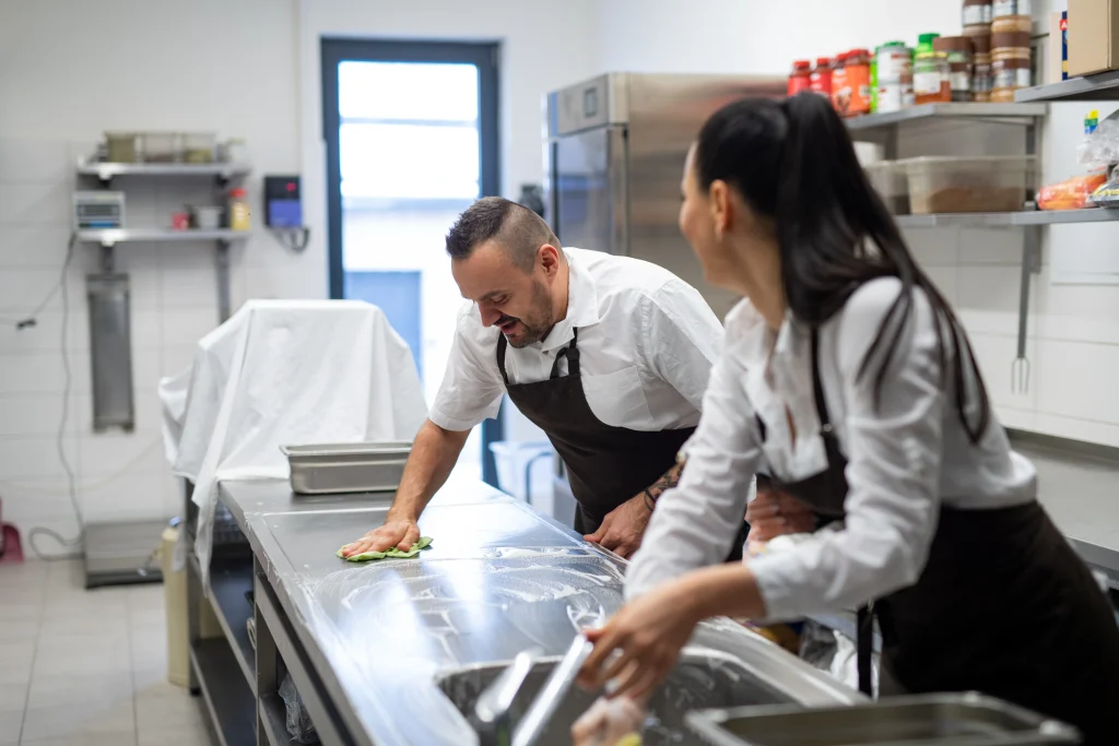 Dois trabalhadores vestindo camisas brancas e aventais escuros higienizam bancada de aço inox em cozinha profissional. Um utiliza pano verde enquanto o outro também limpa a superfície. Ao fundo, prateleiras com recipientes, equipamento coberto e refrigerador reforçam o ambiente de trabalho organizado e limpo.