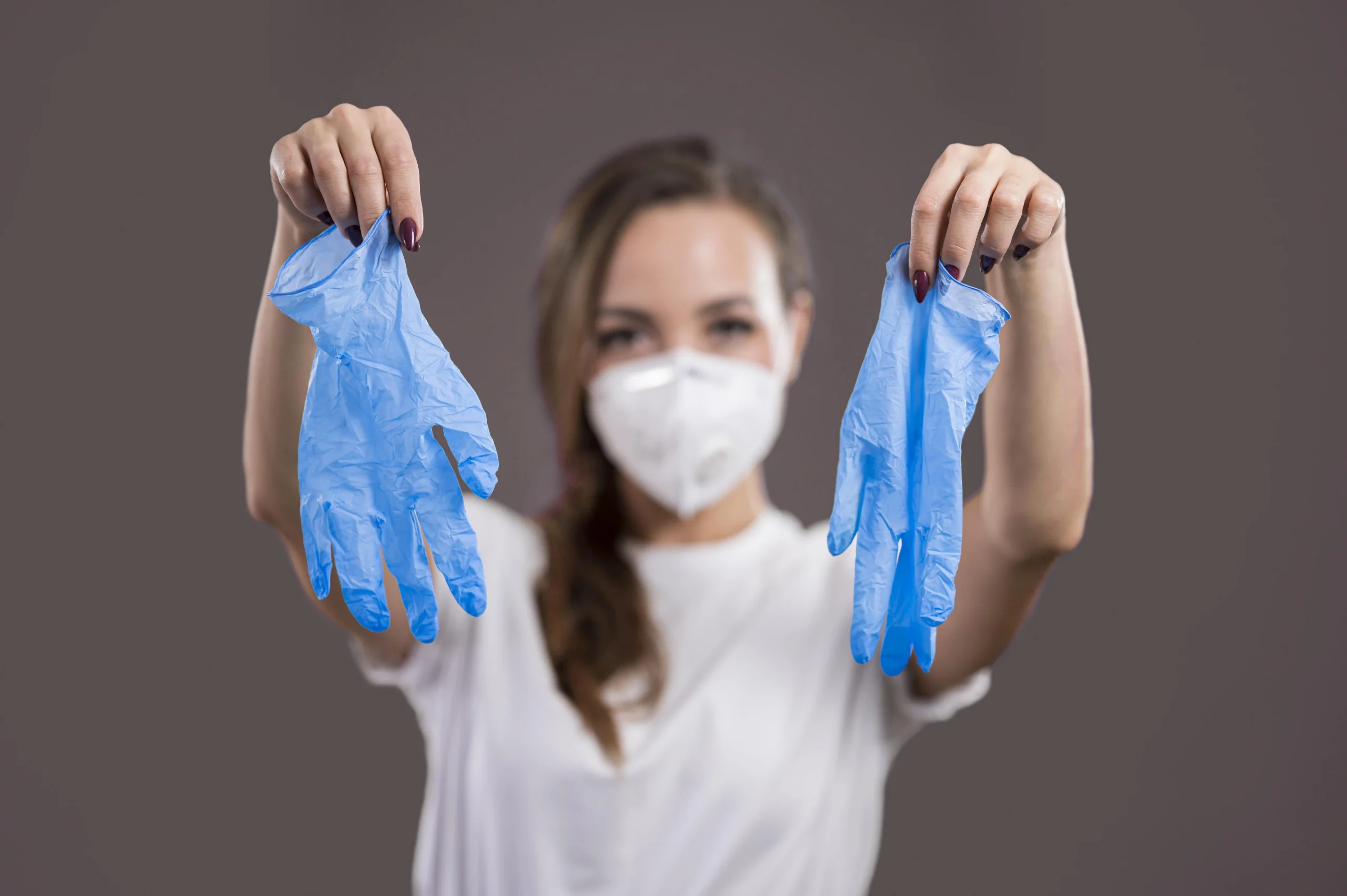 Indivíduo com camisa branca e máscara protetora segura duas luvas azuis descartáveis de látex ou nitrila, uma em cada mão, diante de fundo neutro. A cena enfatiza medidas de segurança, higiene e prevenção de contaminações em contextos médicos ou de saúde pública.