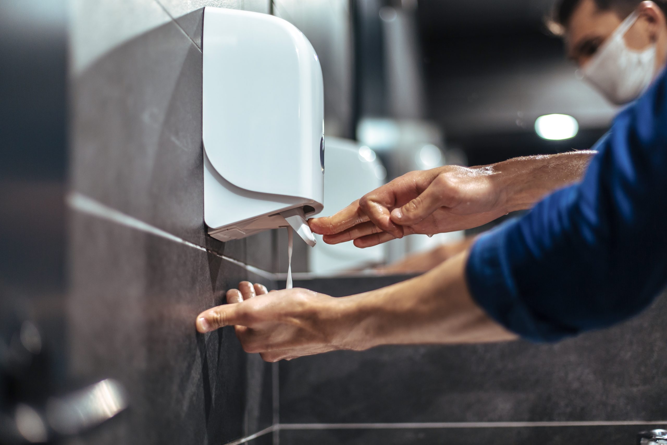 Pessoa usando máscara pressiona dispenser fixado na parede para liberar sabão líquido nas mãos em banheiro com paredes revestidas de azulejos e espelho refletindo a cena. O ambiente reforça práticas de higiene e segurança sanitária em espaços públicos.